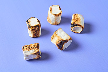 Scattered and grilled white sugary marshmallows cylindrical form lies on purple scratched concrete table. Close-up