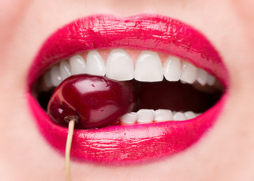 The girl bites a ripe cherry. Cherry in mouth close up. Red lipstick on lips and a cherry with water droplets. The girl is eating cherry.