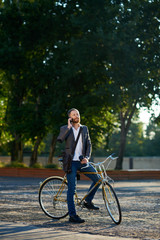 Obraz premium A young stylish bearded businessman in suit going to work by bike. Drinking coffee from a cup to go and talking on a mobile phone on the background of an office building. Takes a working break