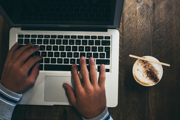 Top view of workstation with modern technology laptop computer and hands to use it and write - coffee or cappuccino drink at its side on wooden table - people at work concept