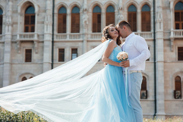 Young beautiful sexy couple hugs and kissing near building, enjoy their romantic date.  Love story in Prague. Woman in blue dress and boyfriend in white shirt. Girl letting her dress flutter in wind