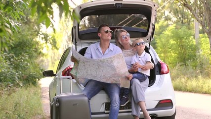 Thoughtful happy parents with son looking at the map sitting in the trunk of the car and dream about traveling. Portrait of a family with a child with a map in the car on the road.