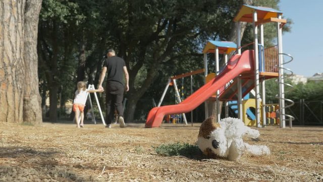 A man in black clothes leads a child out of the playground. Children's soft toy lies on the ground in foreground. Stranger abducted a child