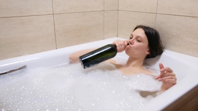 Tired young adult woman drinking wine from bottle lying relaxing in bath tub with foam. Frustrated tipsy person in depression at home in evening. Concept of disappointment in life. Midlife crisis.