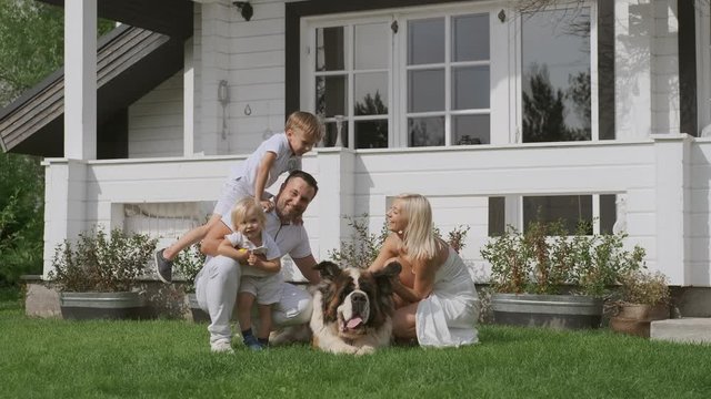 A young family of four in bright clothes and a dog have fun sitting near their white house