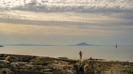 Obraz premium On an early cloudy morning, a fisherman catches fish from a rocky shore. The buoy, which is visible on the right, looks like a big float