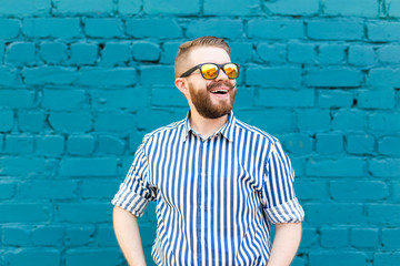 Portrait of a young positive stylish smiling guy with a mustache and beard against the background of a blue brick wall. Vacation concept in the city.