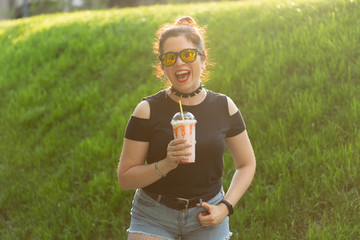 Fototapeta premium Portrait of a positive young pretty girl in punk clothes and glasses with a milkshake in her hands posing in a summer park on a warm summer evening. Vacation getaway concept.