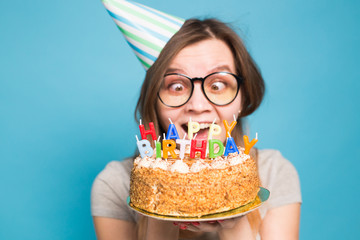 Charming merry crazy young girl student in congratulatory paper hat holding a happy birthday cake in her hands standing against a blue background. Advertising space.