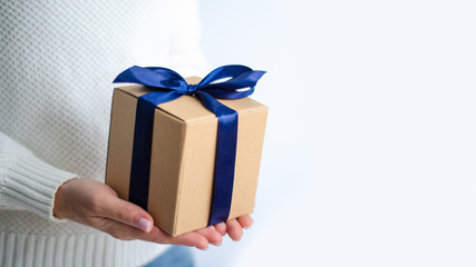 Woman in white sweater holding gift box with blue ribbon in hands on white background