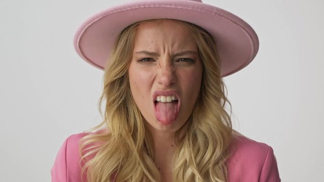 Close up view of unhappy young blonde woman in pink jacket and hat becoming disgusted and showing her tongue while looking at the camera over gray background isolated