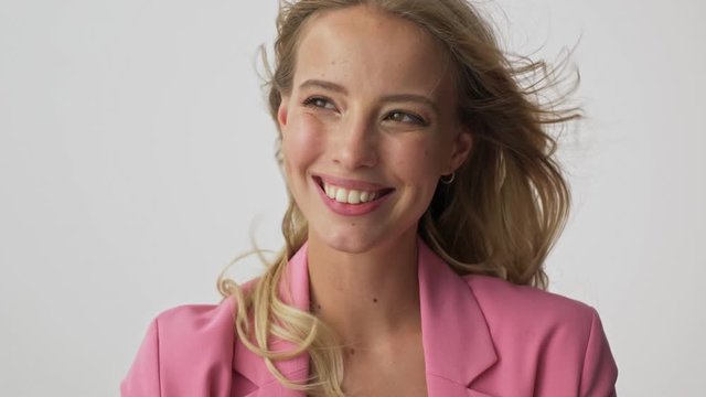 Close up view of happy young blonde woman in pink jacket smiling and touching her fluttering hair while looking at the camera over gray background isolated