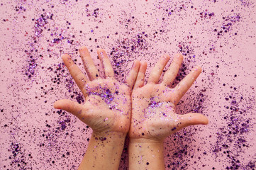 Child's hands with purple color sparkles on pastel pink background.
