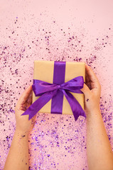 Child's hands holding giftbox tied with purple color ribbon on pink background with glitter. Flat lay style.