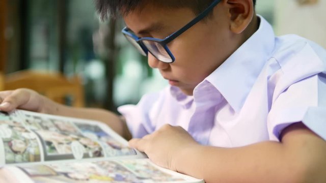 A cute Asian elementary school boy wearing blue glasses in a white school uniform is sitting, enjoying , reading comics in the library.
