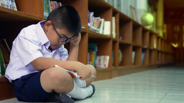 A cute Asian elementary school boy wearing blue glasses in a white school uniform is sitting, enjoying , reading comics in the library.