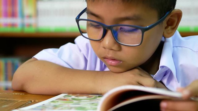 A cute Asian elementary school boy wearing blue glasses in a white school uniform is sitting, enjoying , reading comics in the library.
