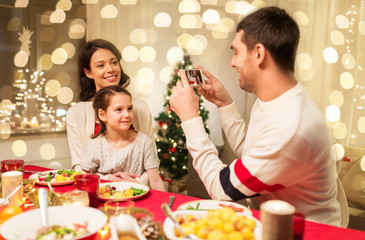 holidays, family and celebration concept - happy mother, father and little daughter having christmas dinner and taking picture by smartphone at home