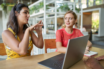 Candid lifestyle shot of two beautiful trendy millennial multi-ethnic students studying together with laptop computer and notepad in bright modern cafe