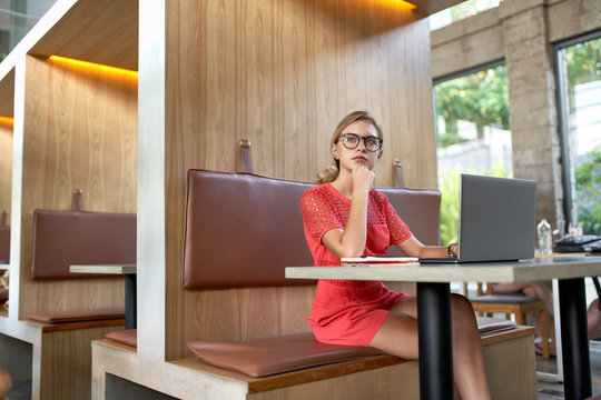 Closeup lifestyle portrait of young pensive blonde caucasian millennial influencer wearing glasses taking notes on a laptop in a bright modern restaurant