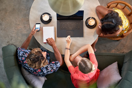 Candid overhead shot of three multi-ethnic millennial coworkers collaborating over coffee with laptop computer at bright cafe serving fair-trade coffee
