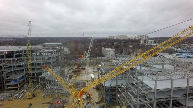 An aerial view of a huge and busy construction site on a puddly ground. Colorful construction cranes are standing between multi storey building frames. The background is filled with leafless forest