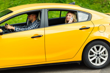 Photo of young woman talking on phone while sitting in back seat of yellow taxi with driver.
