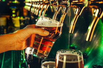 Hand of bartender pouring a large lager beer in tap. Bright and modern neon light, males hands. Pouring beer for client. Side view of young bartender pouring beer while standing at the bar counter.