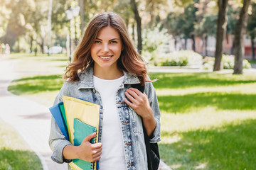 Fototapeta premium Young girl student smiling in the park. Cute girl student holds folders and notebooks in hands, copy space