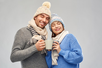 people, christmas and love concept - happy romantic couple in knitted hats and scarves with mugs over grey background