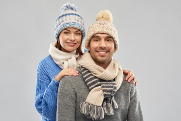 people, christmas and winter clothes concept - happy couple in knitted hats and scarves over grey background