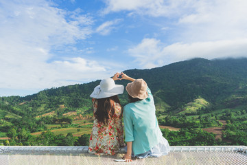 Rear view of couple young Asian woman sitting on the terrace for relaxing and looking mountain and nature view of countryside on morning vacation. Female making heart symbol and shape with hands.