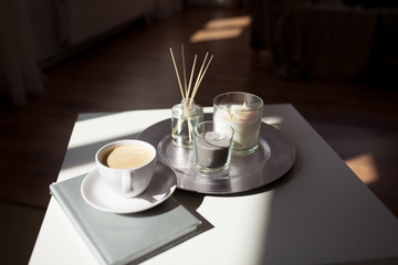 hygge and aromatherapy concept - coffee, candles, book and aroma reed diffuser on table at home