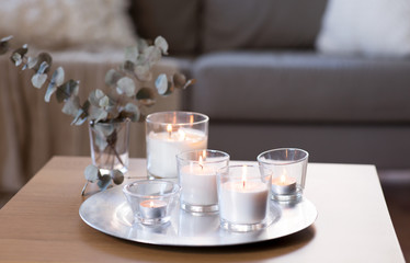 decoration, hygge and cosiness concept - burning white fragrance candle on tray and branches of eucalyptus populus on table at cozy home