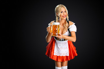 Smiling young sexy oktoberfest girl waitress, wearing a traditional Bavarian or german dirndl, serving big beer mug with drink isolated on black background.