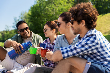 leisure, picnic and people concept - happy friends drinking tea from thermos outdoors in summer park