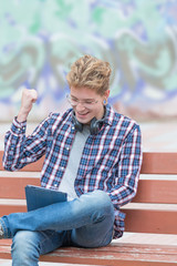 Portrait of a cheerful blond man with glasses and headphones, sitting on a bench reviewing his social networks on a tablet. Concept of technological youth.