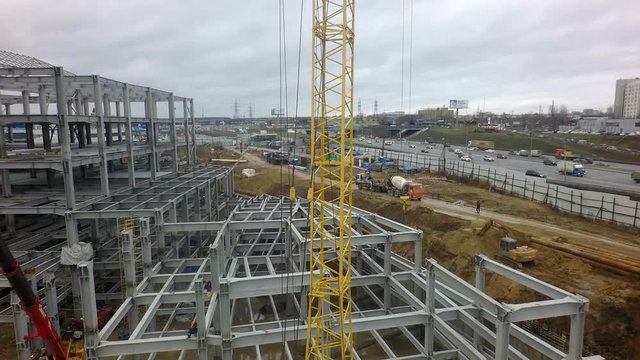 A vertical inspection of a yellow construction crane against the large building frame. Busy workers and construction machines can be seen on the puddly ground of the construction site. A busy highway