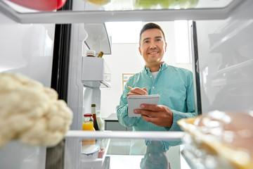 healthy eating and diet concept - smiling middle-aged man opening fridge and making list of...
