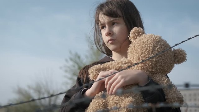Embrace a teddy bear. Homeless child with a toy. Sad girl behind barbed wire with a teddy bear. The child is an orphan.