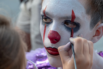 Street makeup in the style of Halloween. A female hand with a brush paints the face of a teenager boy. The image of a clown or mime. All Saints Day celebration or carnival and cosplay competition.