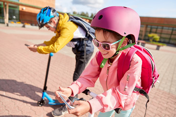 education, childhood and technology concept - happy school children in helmets with smartphones and scooters outdoors