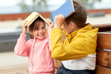 education, childhood and people concept - happy school children or brother and sister with books sitting on wooden street bench outdoors and having fun