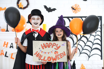 Young girl and boy in costumes with ballons and holding paper with text Happy Halloween