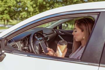 Naklejka premium girl driving car, stands in parking lot, summer waiting for friends and girlfriends, reads message on her smartphone, holds cup of tea her hands with coffee. Summer city background trees.