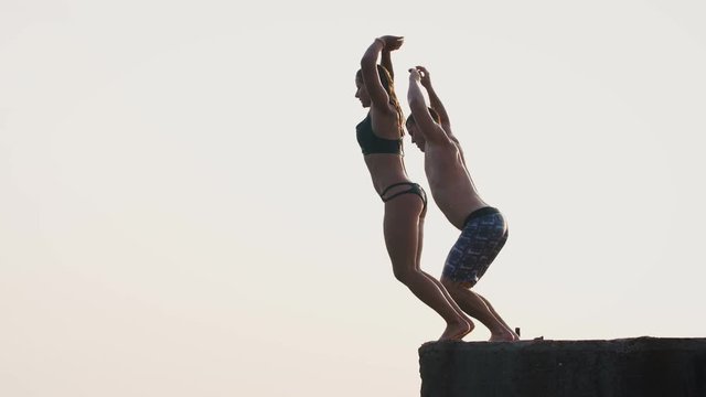 Young woman and man synchronously doing frontflip from a pier into the sea during beautiful sunrise, super slow motion