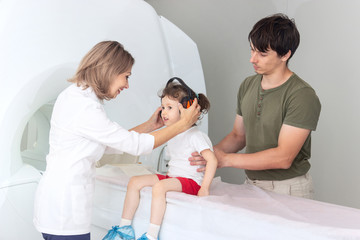 The child sits on a drawer of an MRI machine. Nearby A father and a female doctor are preparing a little girl for magnetic resonance imaging in a hospital. Nurse putting headphones on baby