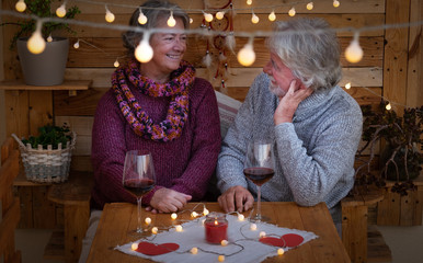 A smiling senior couple looking each other sitting outdoor in a romantic corner made by wood. Yellow lights. Two red wineglasses to celebrate a positive moment