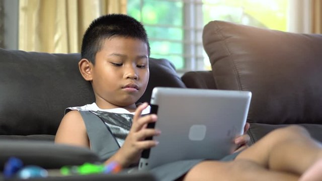 Asian boy playing game or watching cartoons on digital taplet or smartphone on sofa living room.