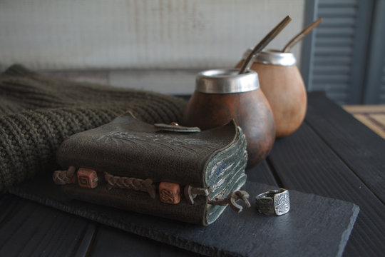 Still life of Two Hand Crafted Artisanal Yerba Mate Tea Leather Calabash Gourd with Straw, leather notebook, sweater and ring on a black painted table, selective focus